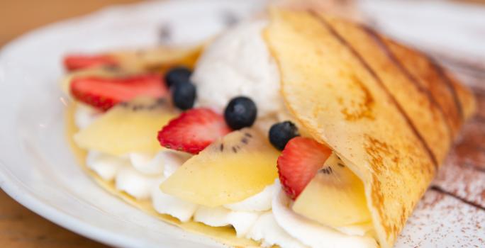 Fotografia em tons de branco com um prato branco ao centro. Em cima do prato existe um crepe recheado com um creme branco doce, morango, kiwi e blueberries.