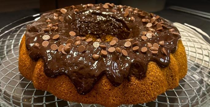 Fotografia de um bolo de cenoura sem glúten sobre um apoio de vidro decorativo. Por cima do bolo com furo no centro tem uma cobertura de chocolate com gotas de chocolate.