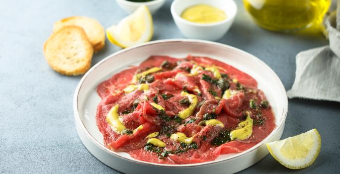 Foto da receita de Carpaccio com Alcaparras e Molho de Mostarda. Observa-se um prato branco grande com a carne fina dentro em rodelas, com molho e alcaparras.