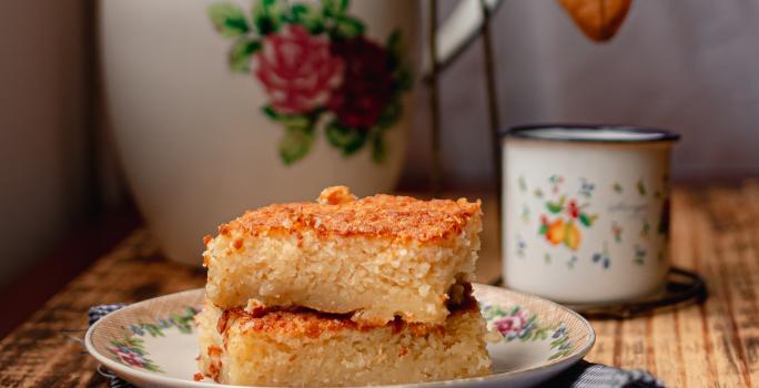 Foto da receita de Bolo de Arroz Cozido. Observa-se 3 fatias retangulares de bolo empilhadas e, atrás, um cafézinho para acompanhar.