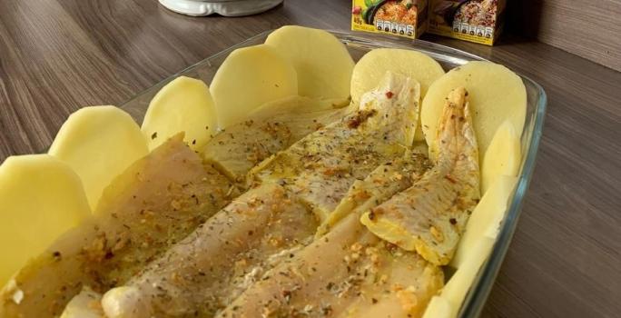 Foto aproximada da receita de Tilápia Assada com Batata no forno, servida em uma travessa de vidro em uma bancada com os temperos maggi meu segredo ao fundo